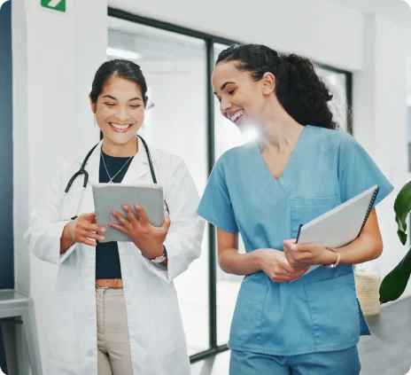 Two women in white and blue uniforms are smiling and holding a tablet.