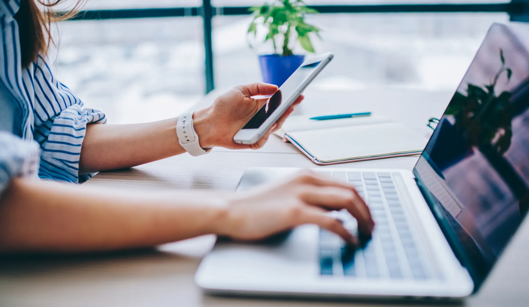 A woman is typing on a laptop with a cell phone in her hand.
