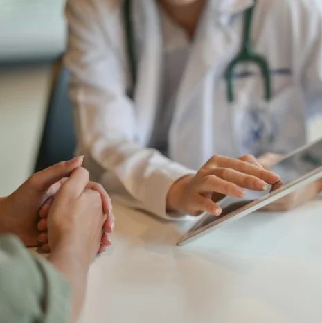 A doctor is using a tablet computer while a patient has their hands clasped together.